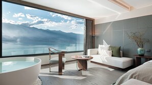 Ein modernes Hotelzimmer mit großem Fenster und Bergblick, einer Sitzecke mit Sofa und Sesseln sowie einem runden Holztisch.