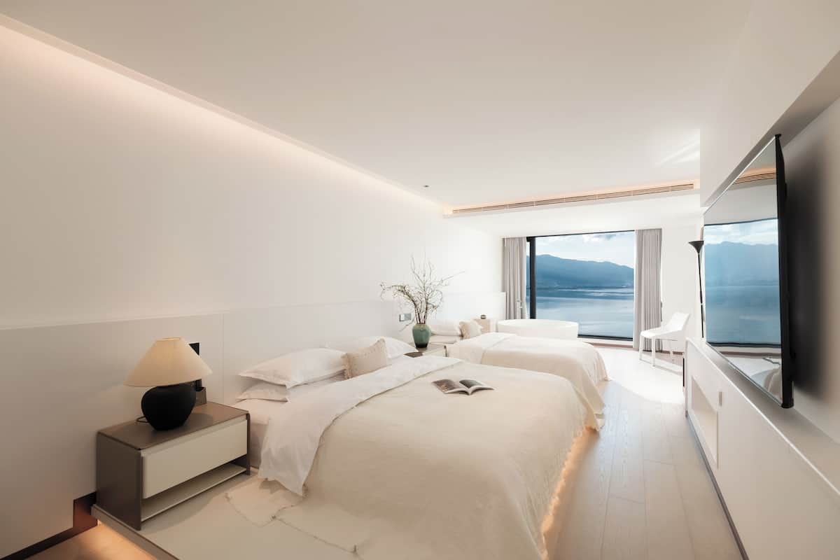Une chambre d’hôtel moderne avec deux lits, une table de chevet avec une lampe, une grande fenêtre donnant sur la mer et un téléviseur à écran plat fixé au mur.