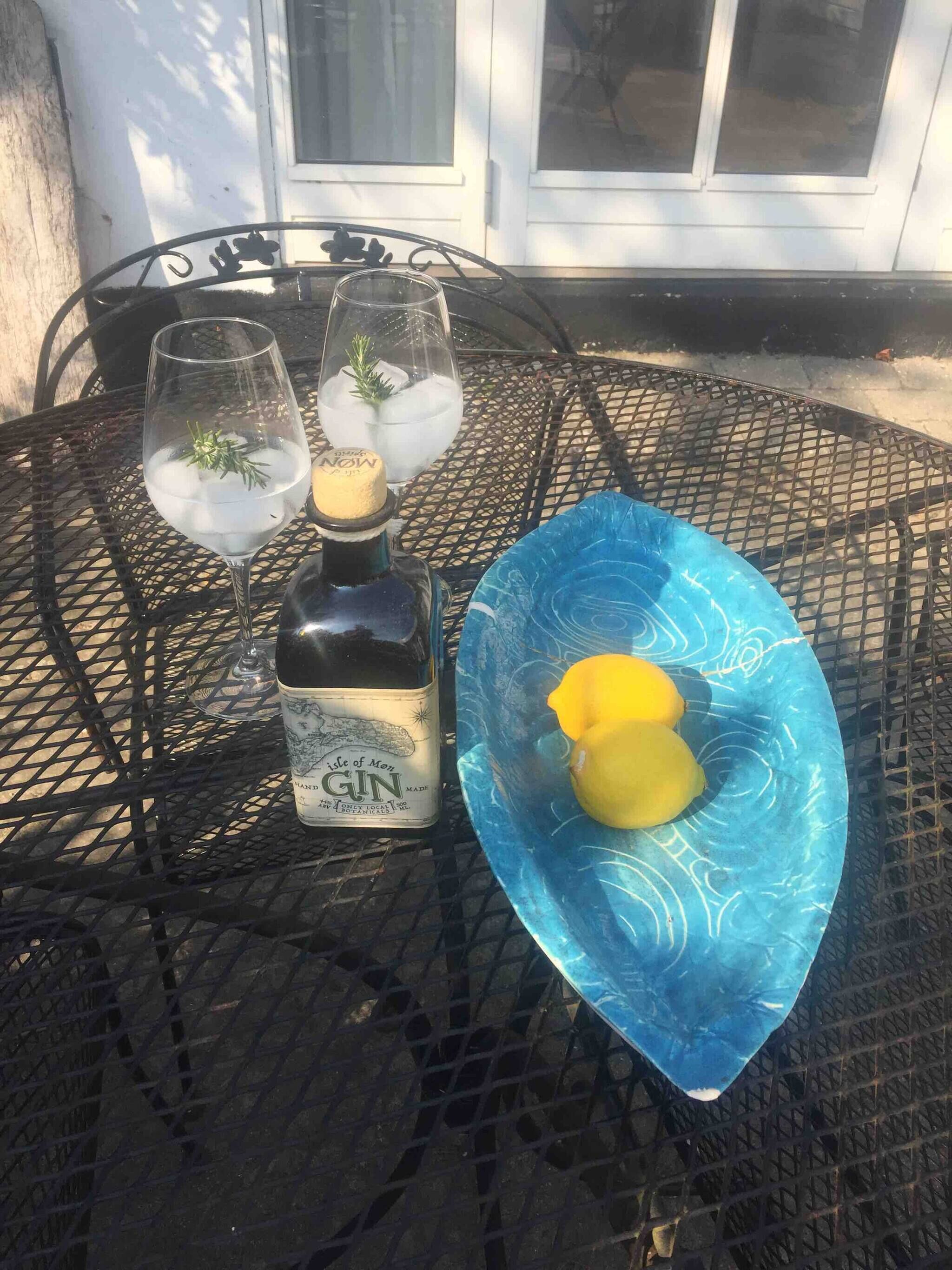 A table with a bottle of gin, two glasses with ice and a sprig of rosemary, and a blue glass bowl with two lemons.