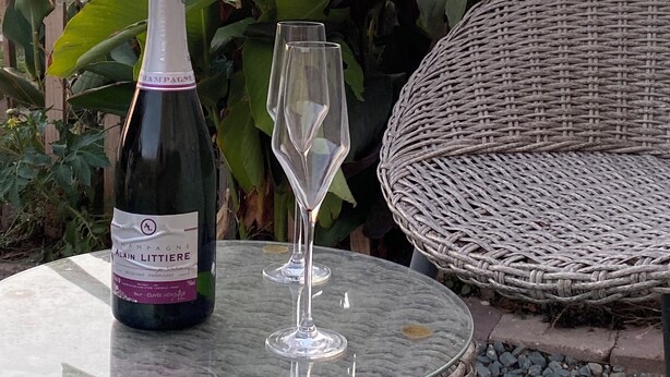 A bottle of champagne and an empty glass on a round glass table outdoors with a wicker chair and large green plants in the background. - Between land and sea (Marennes-Hiers-Brouage)