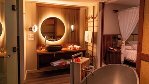 A hotel bathroom with a bathtub, a sink with a round mirror, and a vanity with toiletries. - HOTEL WADRA BAY (Lifou)