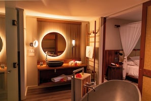 Une salle de bain d’hôtel avec une baignoire, un lavabo avec un miroir rond et une commode contenant des articles de toilette.