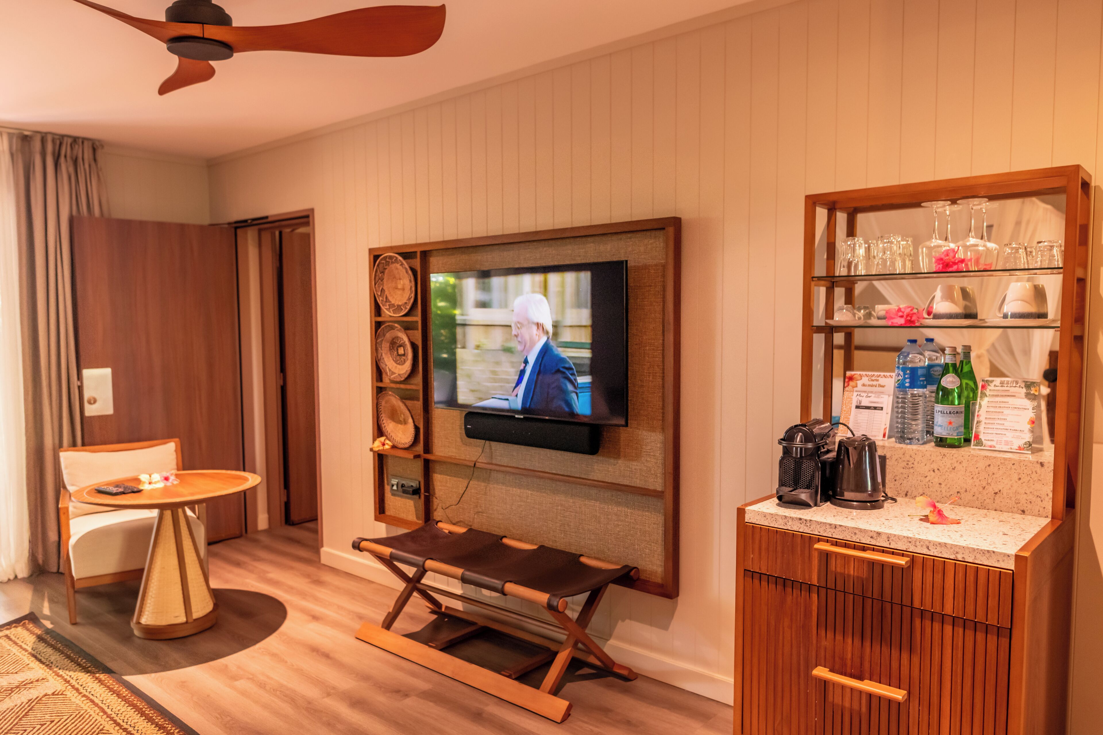 A hotel room with a television mounted on the wall, a small table with a chair, a bar area with glasses and bottles, and a wooden floor.