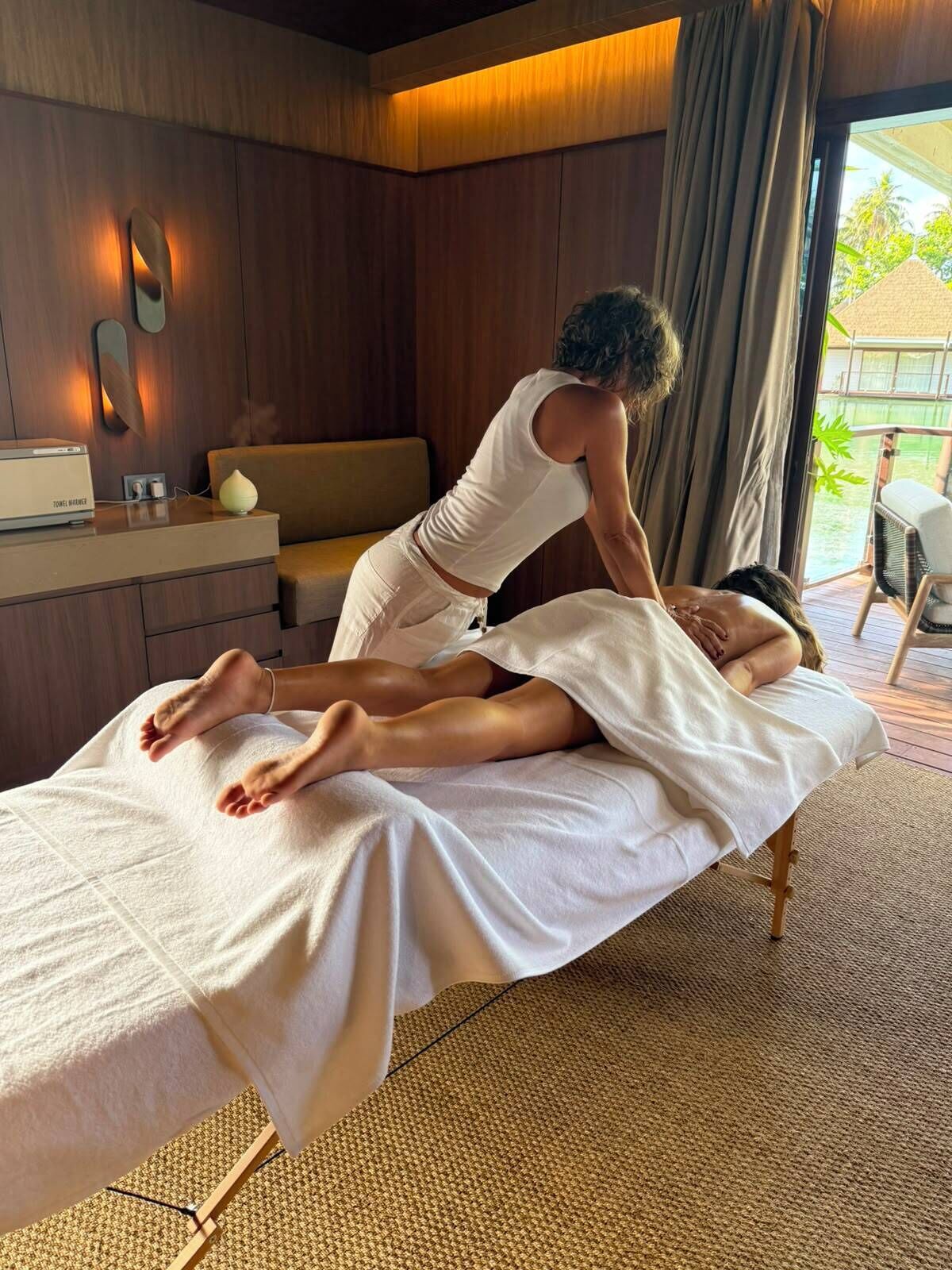 Een massage therapie sessie in een ruimte met een houten wand, een massagetafel, een kleine bank, een bijzettafeltje met een lamp, en uitzicht op het buitenlandschap door een raam.