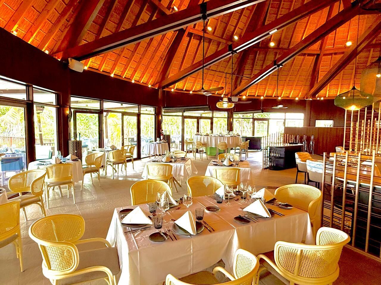 A restaurant with multiple tables and chairs, large windows, and a high ceiling with wooden beams.