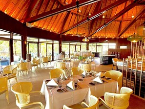 A restaurant with multiple tables and chairs, large windows, and a high ceiling with wooden beams. - HOTEL WADRA BAY (Lifou)