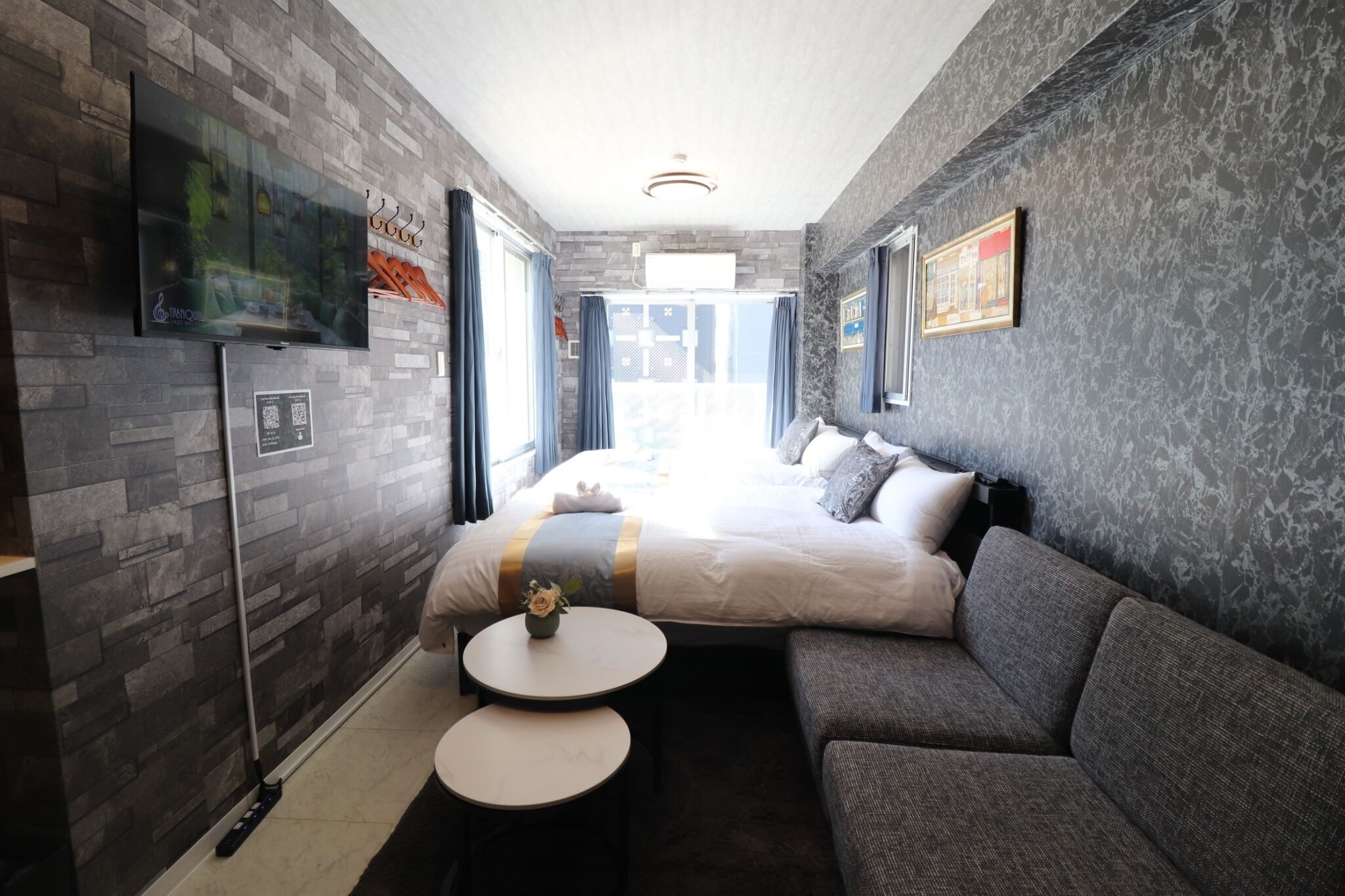 A modern hotel room with a large bed, a sofa, two small tables, a TV, and a view of the outside.