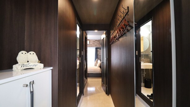 A modern hallway with dark wood paneling, a white reception desk with a 'Welcome' sign, and a view into a bedroom with a bed and a bathroom visible through a glass door. - One Stay Shinsaibashi (Osaka)