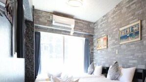 A hotel room with two beds, a round table, a small sofa, a window with curtains, and a wall-mounted air conditioner. - One Stay Shinsaibashi (Osaka)