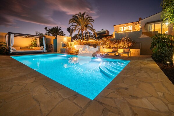 A swimming pool with a canopy and lounge chairs, surrounded by palm trees and a modern building.