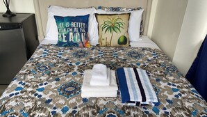 Un letto rifatto con cura, con copriletto decorato, due cuscini con scritte e un motivo di palma, un asciugamano bianco piegato e un asciugamano a righe.
