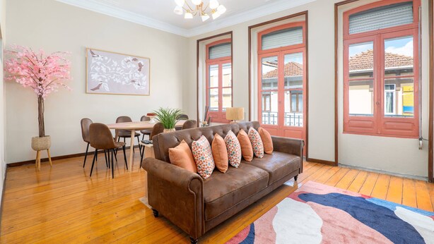 Ein Wohnzimmer mit einem braunen Sofa, einem Esstisch, Stühlen, einer Vase mit rosa Blumen und großen Fenstern mit roten Rahmen.