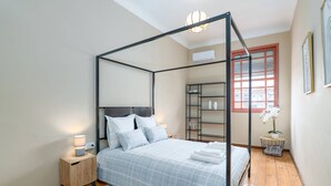 Ein Bett mit Baldachin, dessen Bettdecke weiß-blau kariert ist, ein Nachttisch mit Lampe und ein kleiner Topfpflanze auf einem runden Tisch neben dem Fenster.
