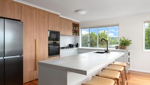 Una cocina moderna con un refrigerador, horno y microondas de acero inoxidable, armarios de madera, una isla grande con taburetes de bar y una ventana con vistas a palmeras.