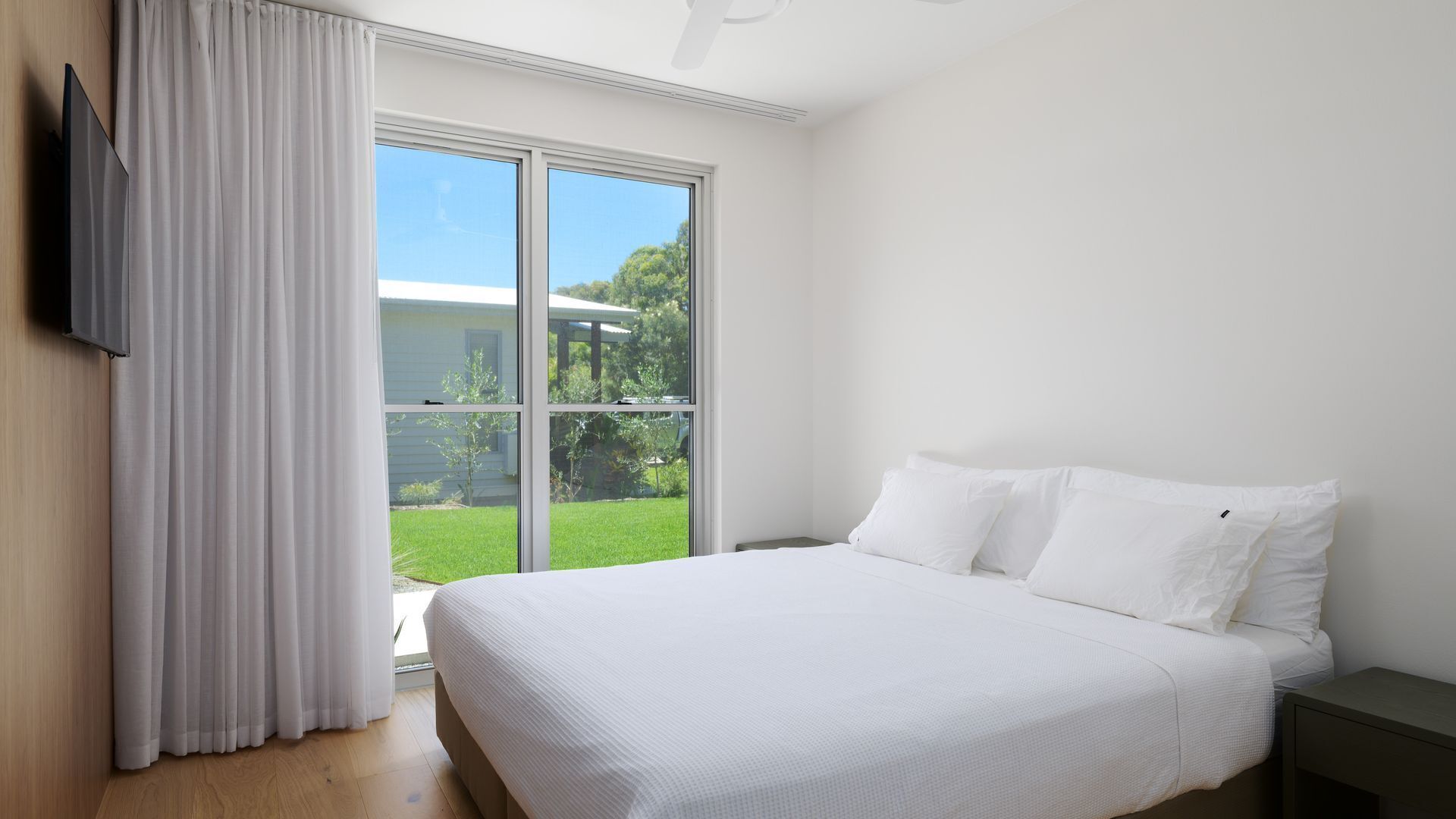 A bedroom with a large bed, a TV mounted on the wall, and a sliding glass door leading to a garden.