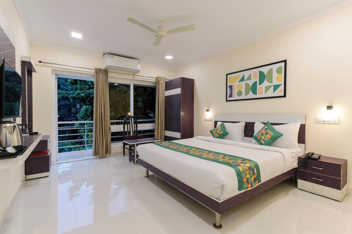 A hotel room with a large bed, a desk with a kettle, a chair, a balcony with a view of trees, and a ceiling fan.