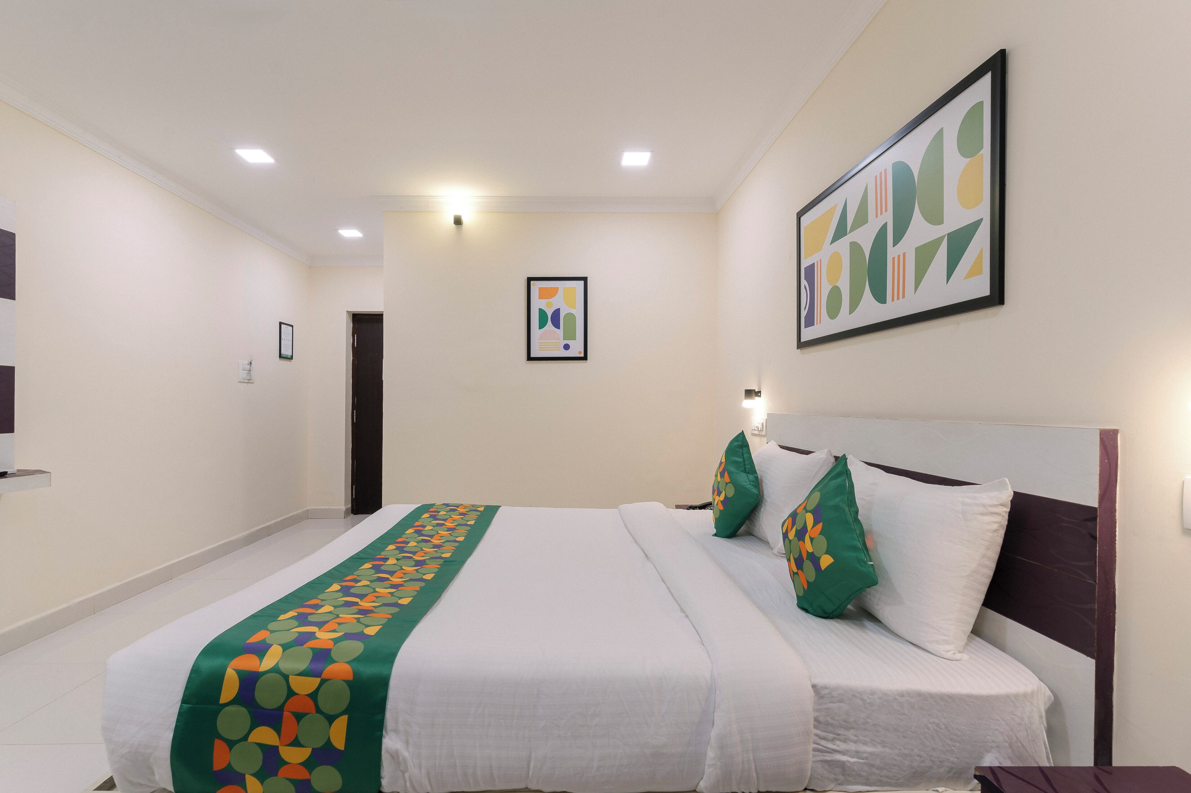 A hotel room with a large bed, a green patterned bedspread, white pillows, and a framed abstract artwork on the wall.