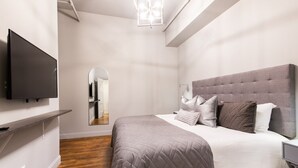 A modern bedroom with a large bed, a flat-screen TV mounted on the wall, and a mirror reflecting the room. - Hollyhock Suite 503: 2 bed/1 bath (Louisville)