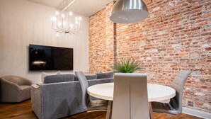 A modern living room with a brick wall, a round dining table, grey sofas, and a large TV. - Hollyhock Suite 503: 2 bed/1 bath (Louisville)