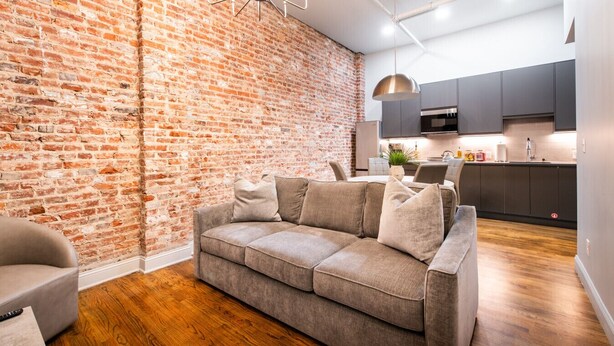 A modern living room with a brick wall, a grey sofa, wooden flooring, and a kitchen area with dark cabinets and a stainless steel microwave. - Hollyhock Suite 503: 2 bed/1 bath (Louisville)