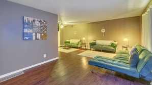 Una sala de estar con un sofá azul, sillones verdes y una mesa de centro. Hay un cuadro en la pared y una mesita con una lámpara.