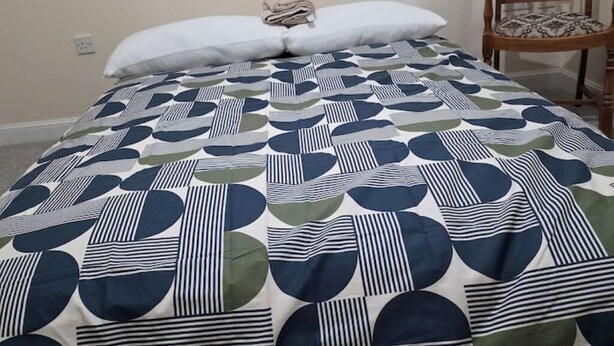 Ein Bett mit einem geometrisch gemusterten Überwurf, einem weißen Kopfkissen und einem Holzstuhl mit einem gemusterten Sitzkissen.