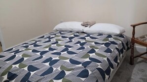 Ein Einzelbett mit einem gemusterten Bettbezug, ein Holzstuhl und eine schlichte Wand.