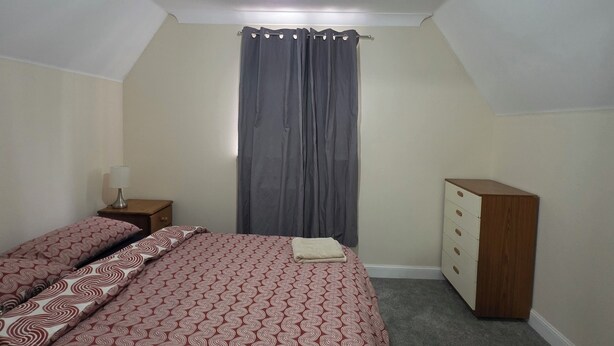 A bedroom with two beds, a dresser, a nightstand, a lamp, and a window with curtains. - Stay Near Colchester twn centre4 (Colchester)