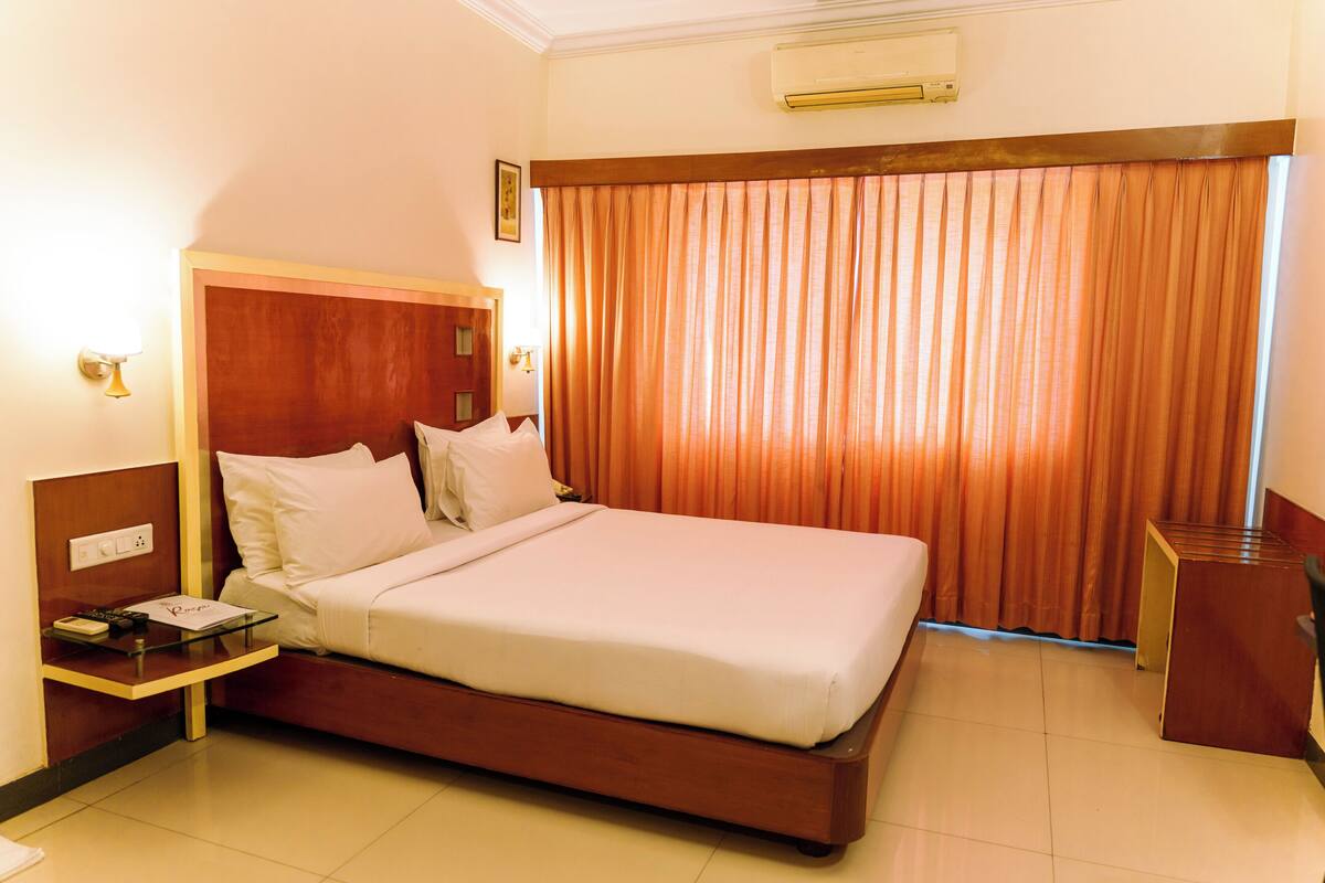 Habitación de hotel con una cama de madera, una mesita de noche con lámpara y un ventanal con cortinas.