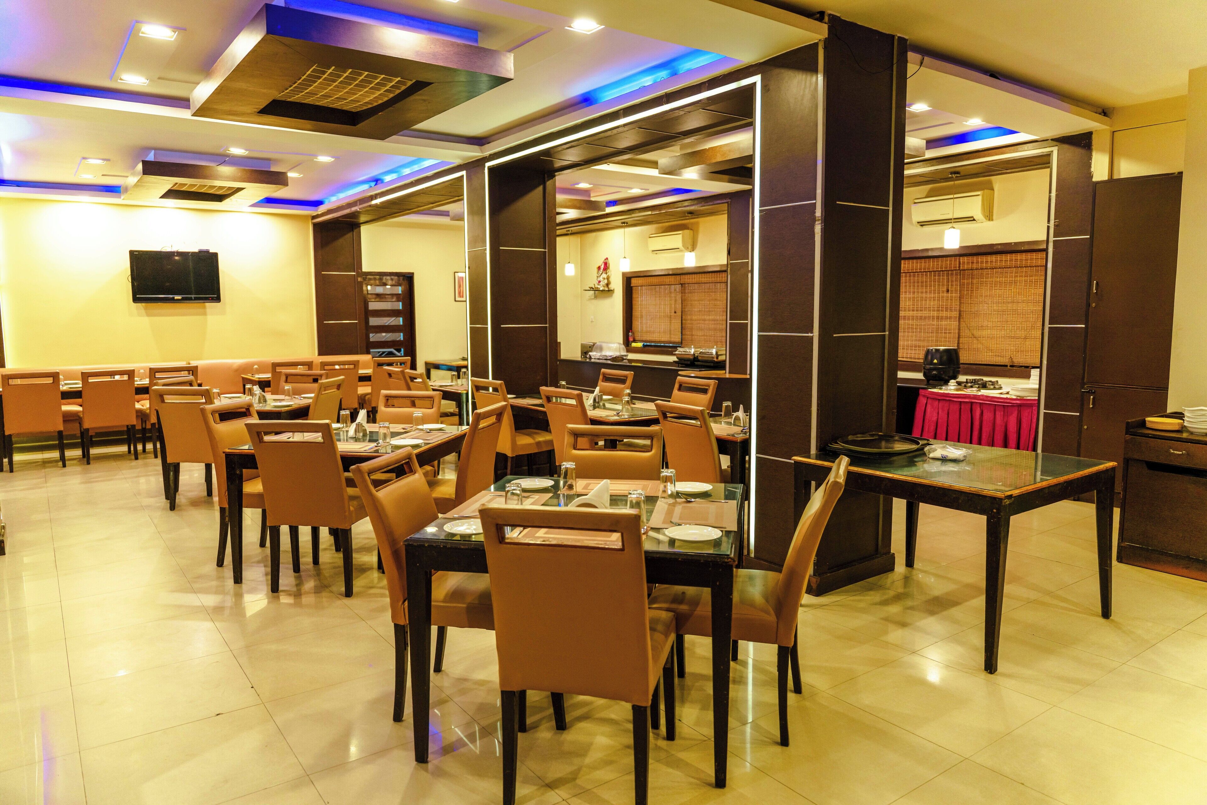 A restaurant with multiple tables and chairs, a television mounted on the wall, and a kitchen area visible through a partition.
