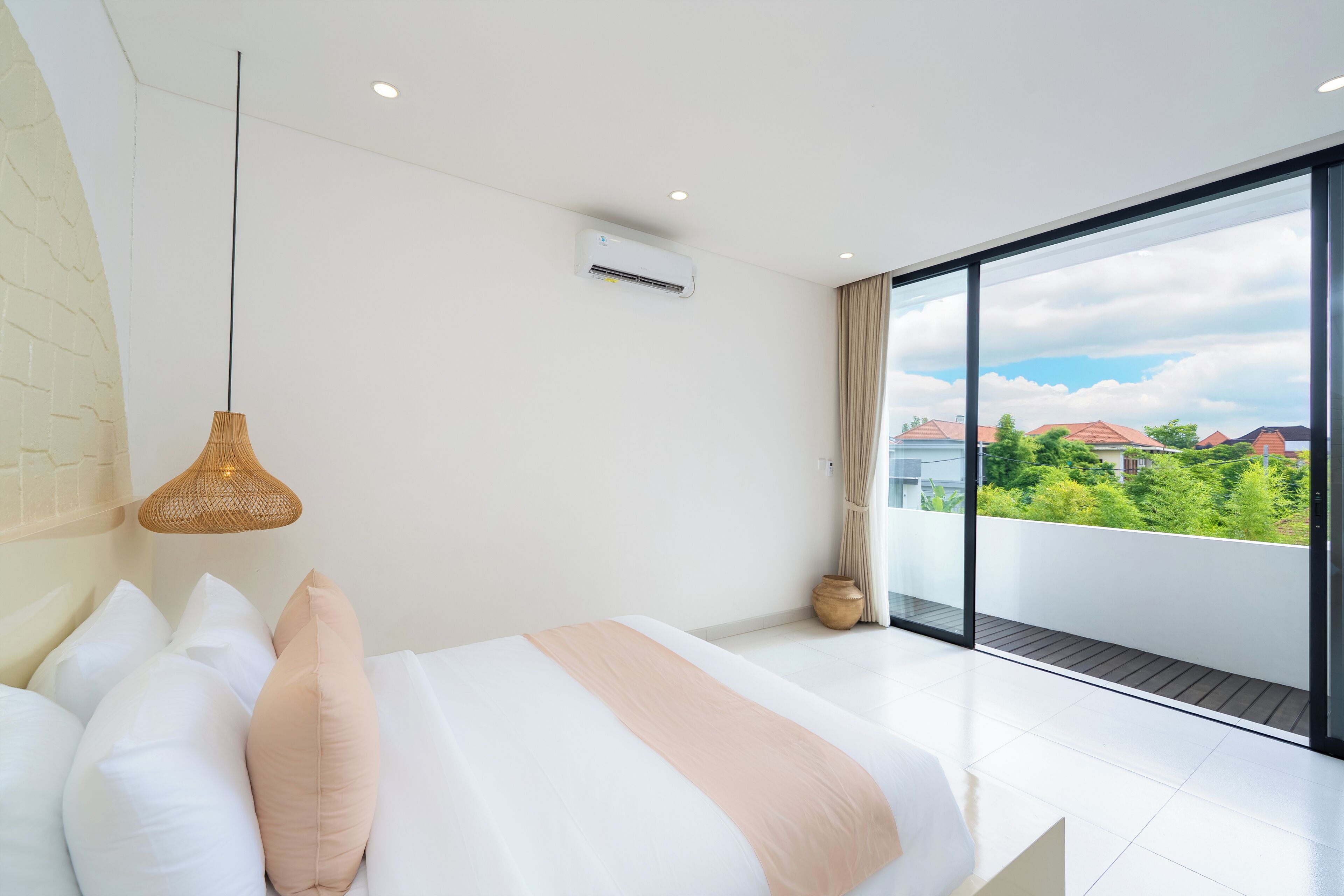 A modern bedroom with a large bed, a hanging light fixture, and a view of greenery through large windows.
