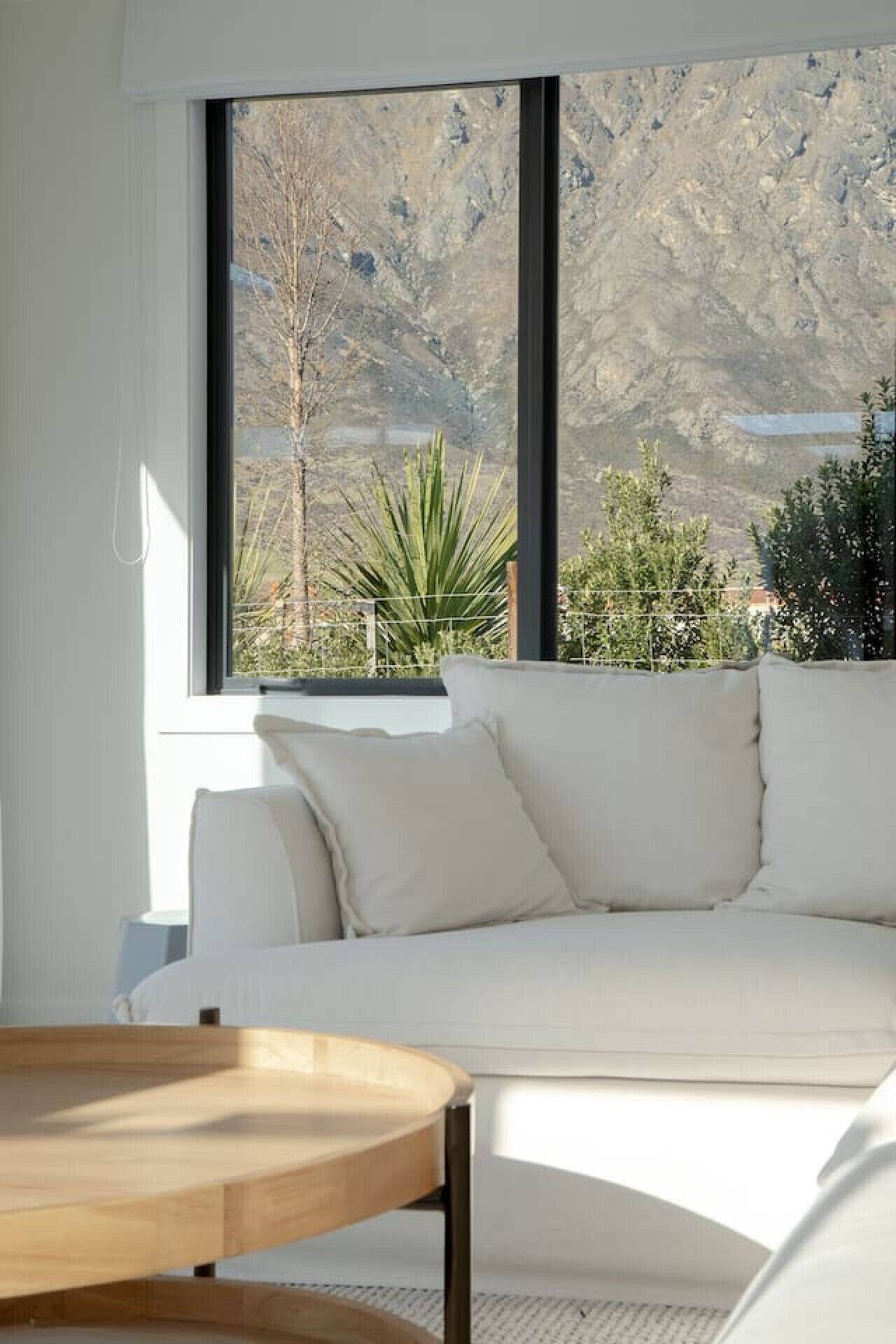 A modern living room with a white sofa, a wooden coffee table, and a large window offering a view of a mountainous landscape.