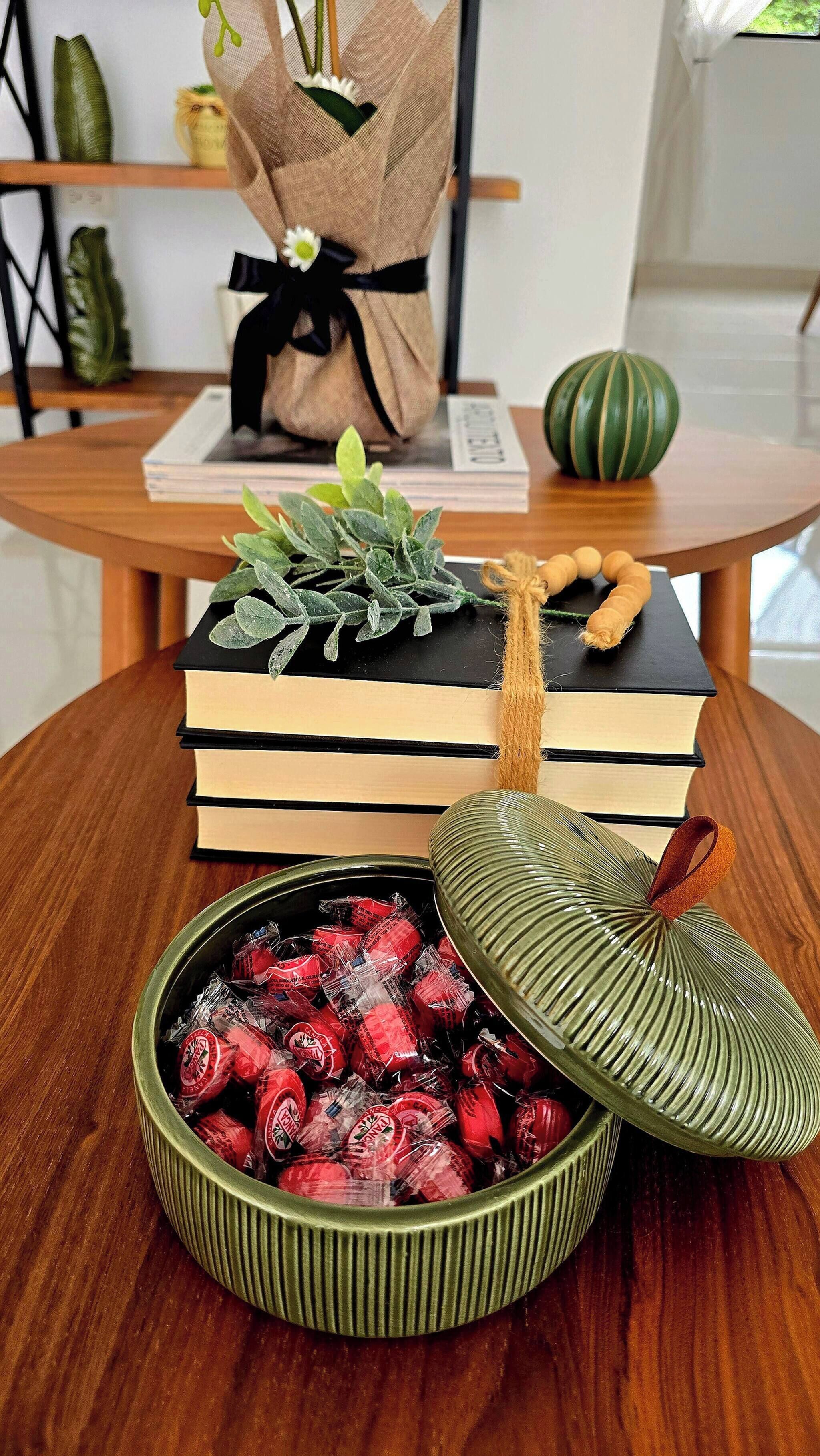 Uma mesa de madeira com uma pilha de livros, uma pequena planta, uma tigela decorativa com balas e um enfeite verde.