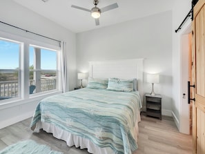 A bedroom with a bed, two bedside tables, a ceiling fan, and a view through a large window.