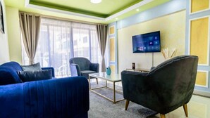 Een moderne woonkamer met een blauwe bank, een zwarte fauteuil en een glazen salontafel. Een flatscreen televisie is aan de muur bevestigd, en er is een raam met gordijnen.