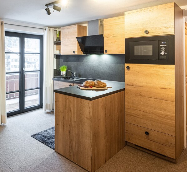 A modern kitchen with wooden cabinets, a black microwave, and a small island with a cutting board and bread.