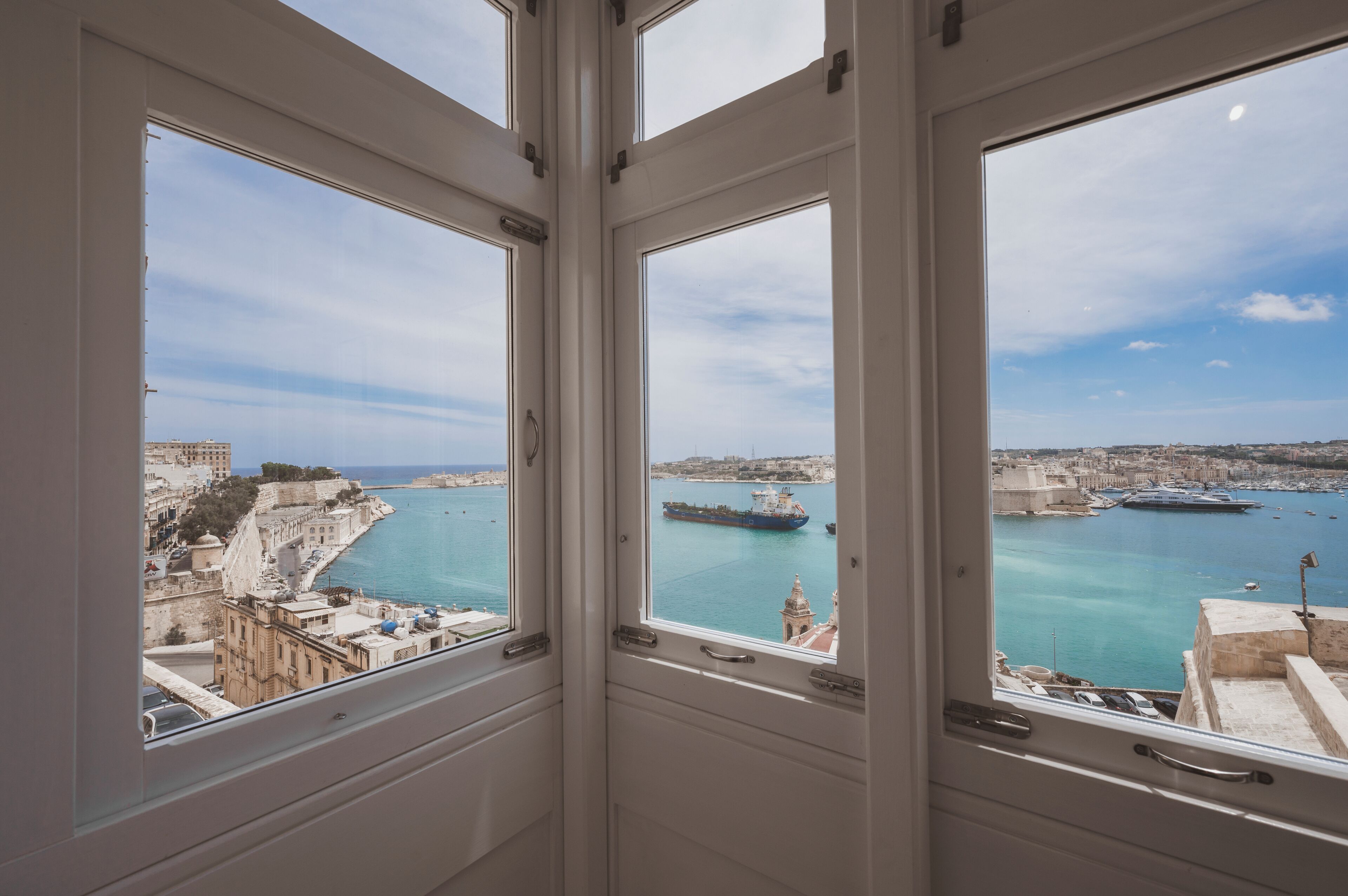 Una stanza con ampie finestre che si affaccia su una città costiera, con un porto, imbarcazioni ed edifici storici.