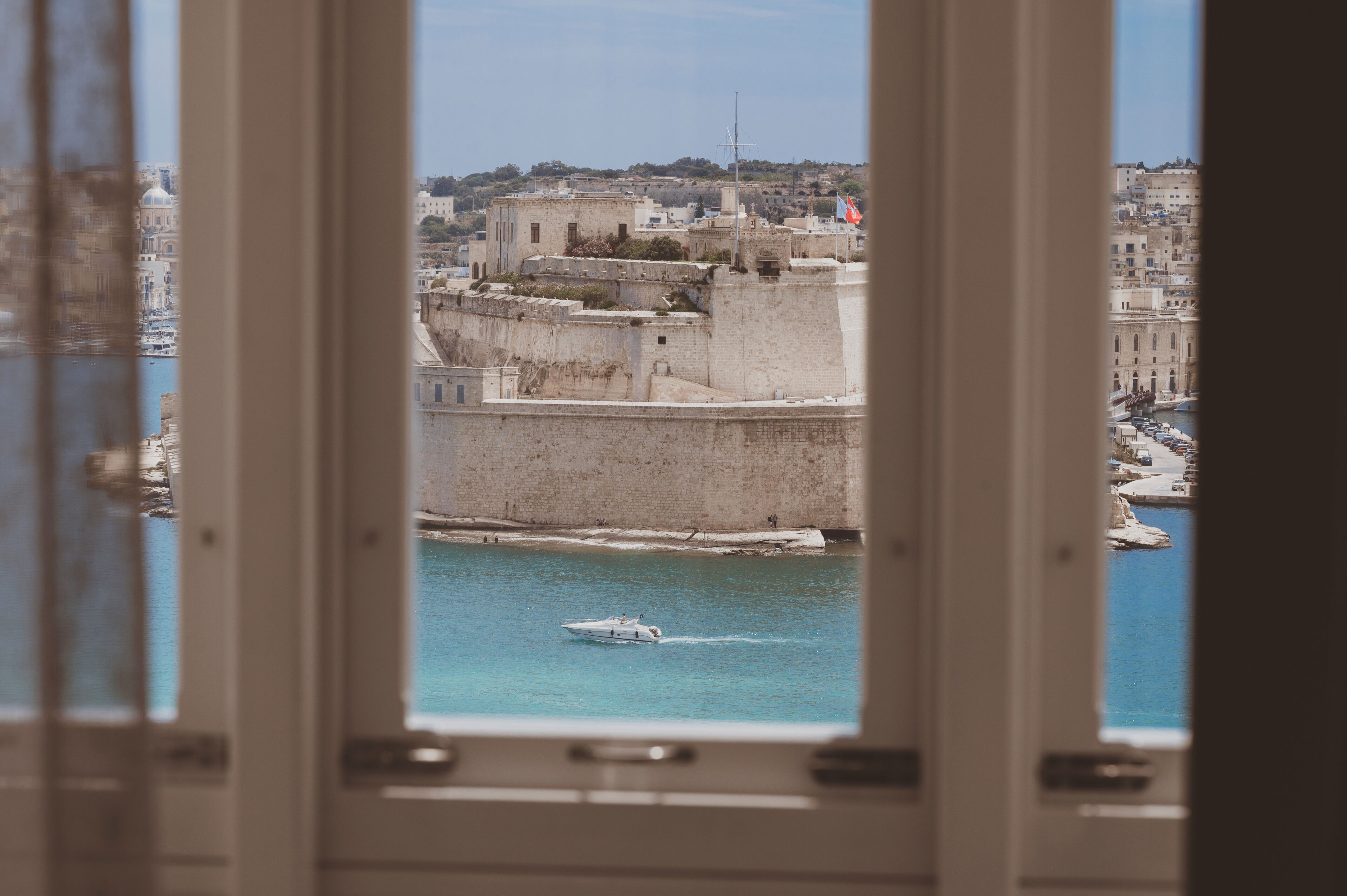 Vista de una ciudad costera a través de una ventana, con una fortaleza histórica, una masa de agua y un barco.