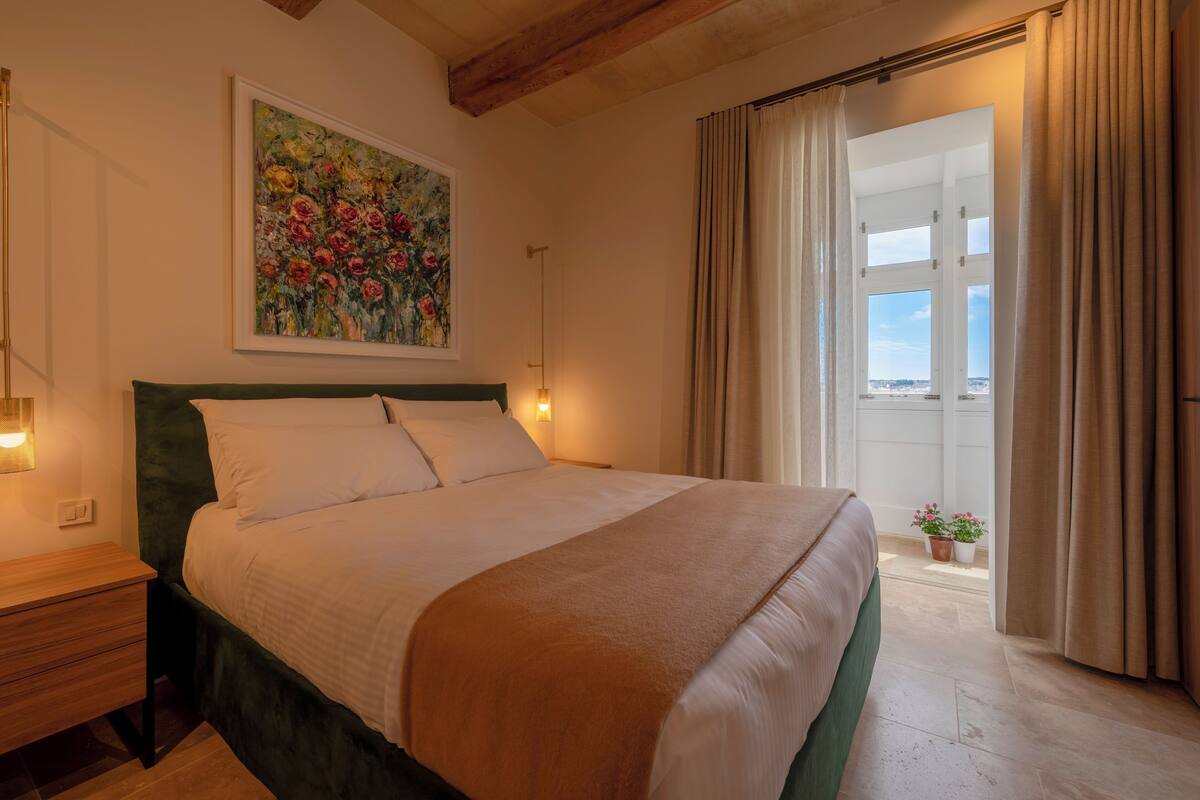 Una cama bien hecha con cabecera verde, un cuadro encima y vista al exterior a través de una ventana con cortinas.