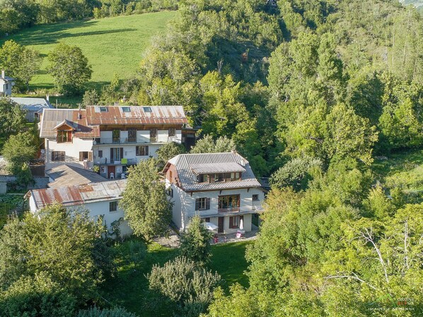 Une grande maison dotée d’un balcon, entourée d’arbres denses et d’un champ verdoyant.