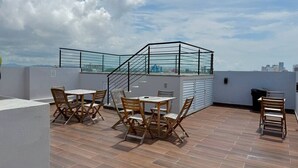 A rooftop terrace with wooden tables and chairs, a glass railing, and a city view.