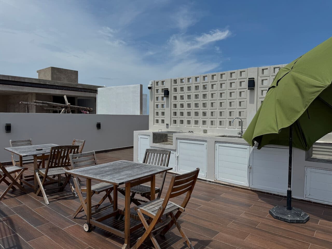 Une terrasse extérieure avec des tables et des chaises en bois, un parasol vert et, en arrière-plan, un bâtiment blanc dont la façade est ornée d’un motif en forme de quadrillage.