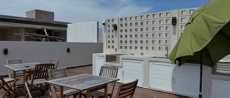 Une terrasse extérieure avec des tables et des chaises en bois, un parasol vert et, en arrière-plan, un bâtiment blanc dont la façade est ornée d’un motif en forme de quadrillage.