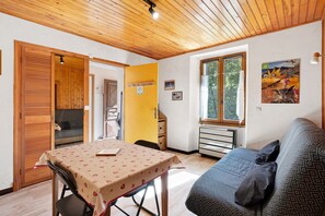 A cozy room with a wooden ceiling, a dining table with a tablecloth, a sofa, and a window with a view of greenery.