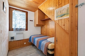 A small, cozy room with wooden walls and a sloped ceiling. There is a bed with a striped blanket and a window with a curtain.