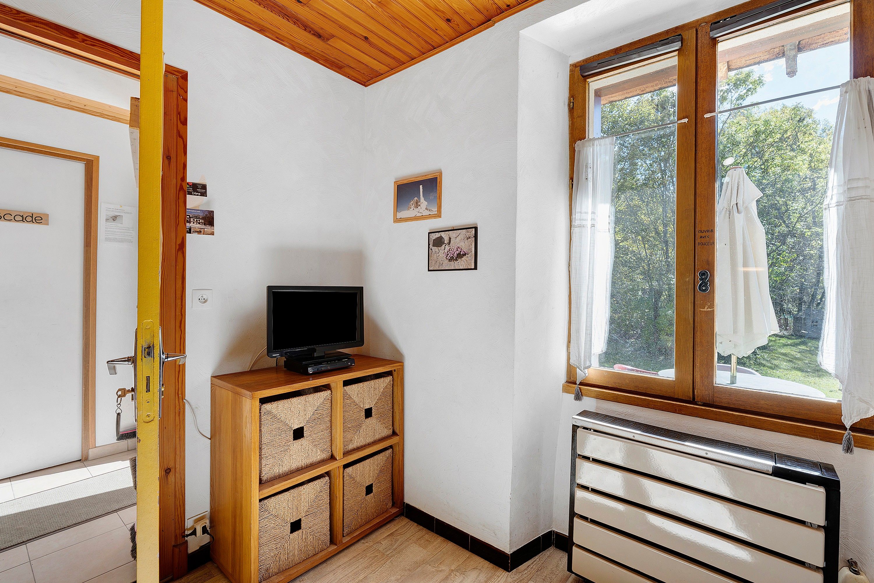 Une pièce avec un plafond en bois, un téléviseur posé sur un meuble en bois, une fenêtre donnant sur des arbres et un radiateur.