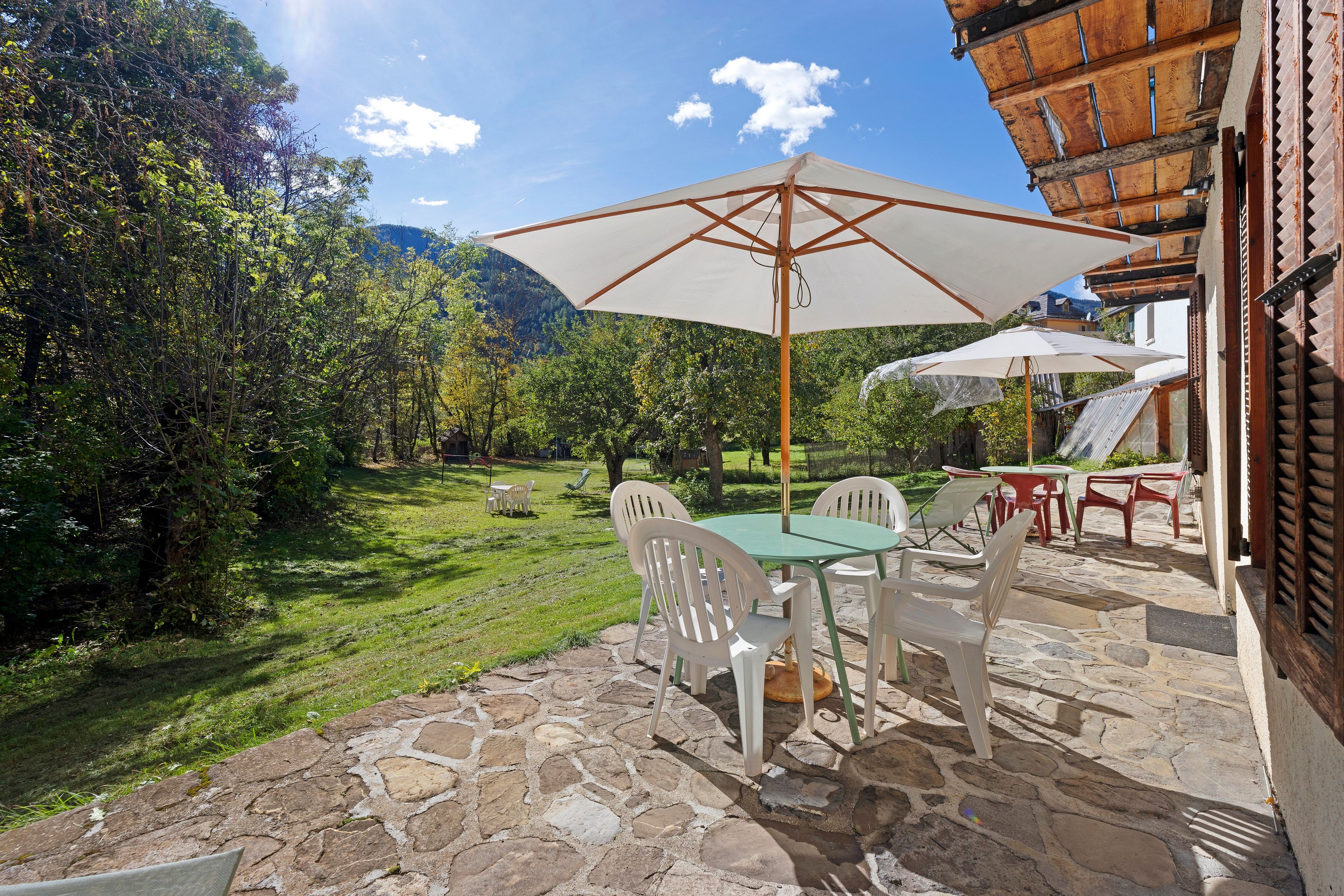 Une terrasse extérieure avec un sol en pierre, des chaises en plastique blanches et une table placée sous un grand parasol.