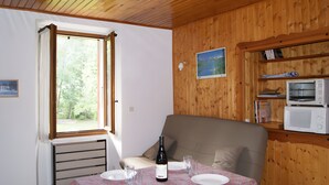 A room with a dining table set for two, a sofa, a window with curtains, a radiator, a microwave, and a shelf with books. - Gite la petite cascade - Haut Verdon Val d'Allos (Allos)
