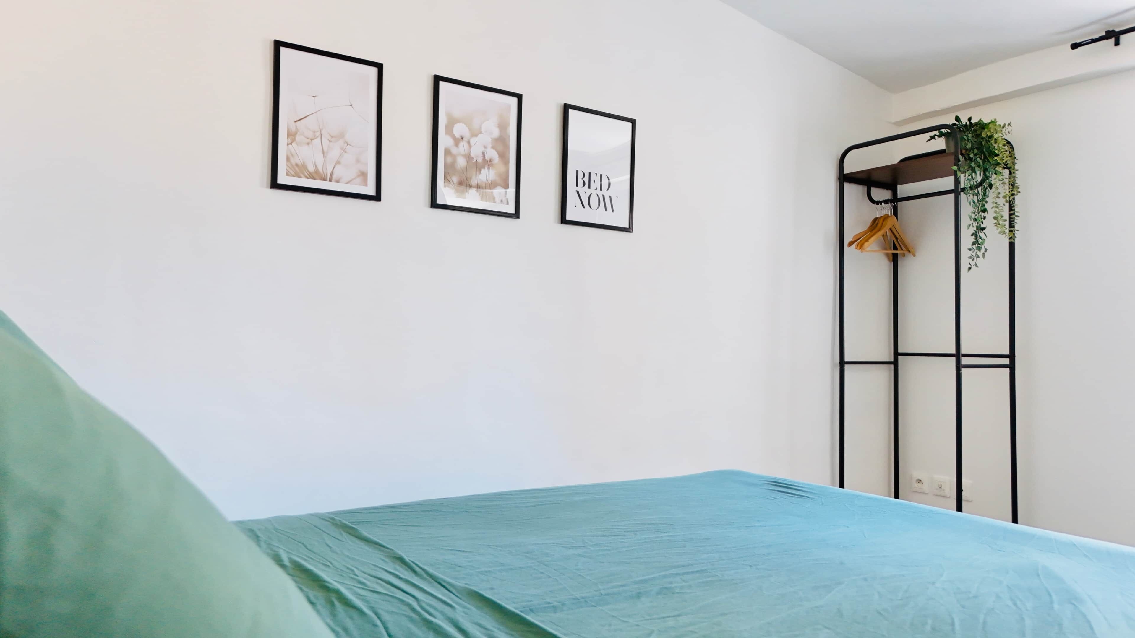 Une chambre à coucher avec un lit, une étagère fixée au mur ornée d’une plante, et trois tableaux encadrés.