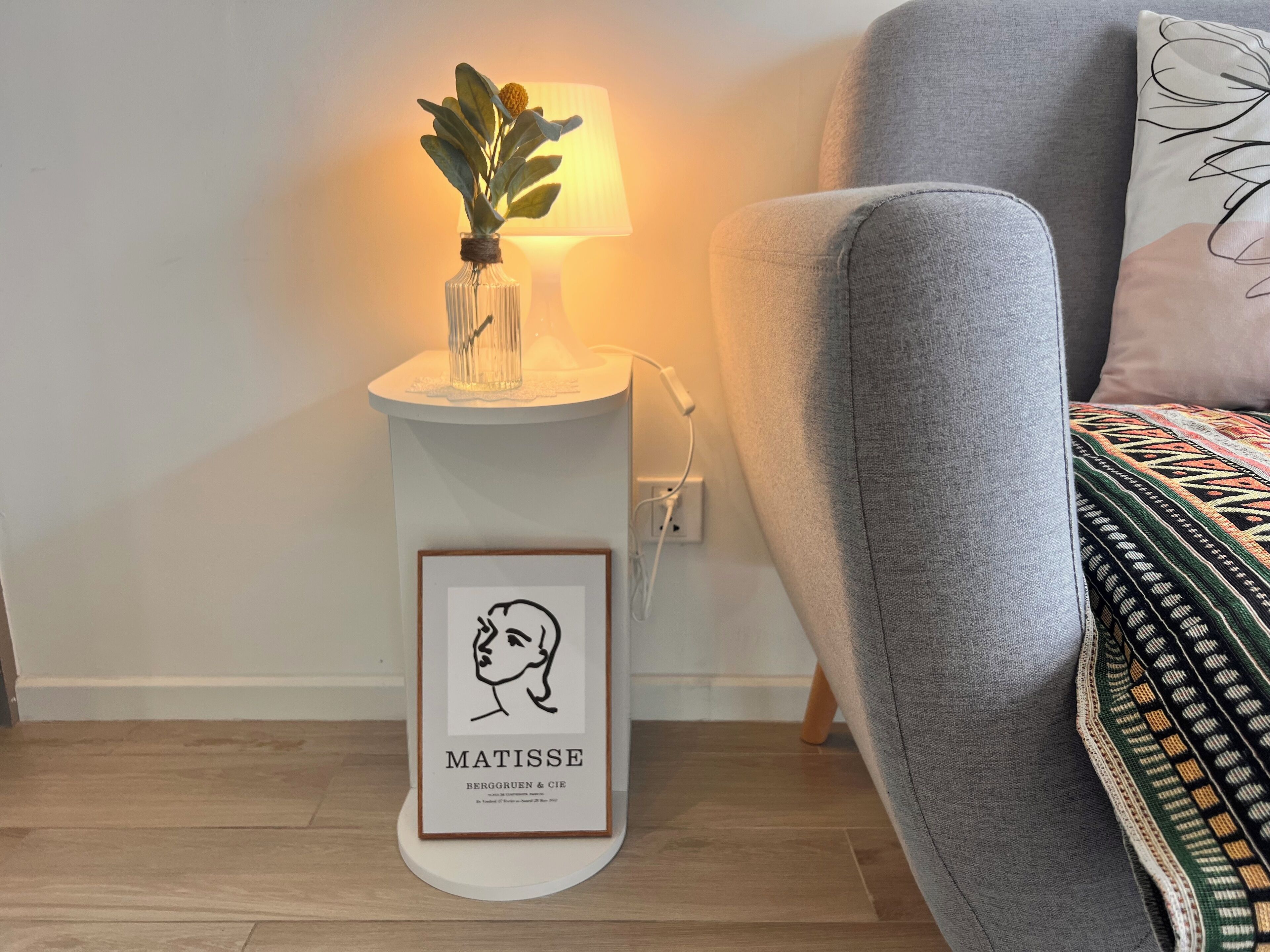 Una mesita con una obra enmarcada de un rostro, un pequeño florero con una planta y una lámpara.